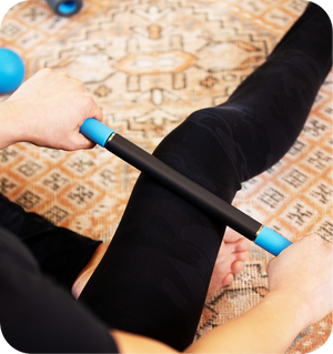 Person using a RAD ROD | RAD massage tool on their calf while sitting on a patterned carpet, promoting muscle recovery and wellness.