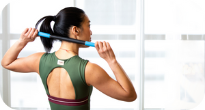 Woman in green sportswear using a RAD ROD | RAD wellness recovery tool to massage her upper back indoors by a large window.