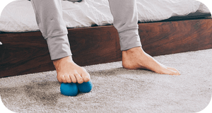 Person using blue massage roller under foot on carpet near wooden bed frame featuring RAD Roller Stiff | RAD wellness recovery tool