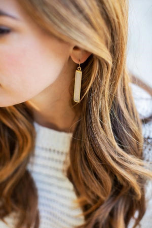 Close-up of a woman wearing Selenite Earrings | Meghan Bo that feature a rectangular selenite stone set in a gold-colored frame, complementing her wavy light brown hair and cream sweater.