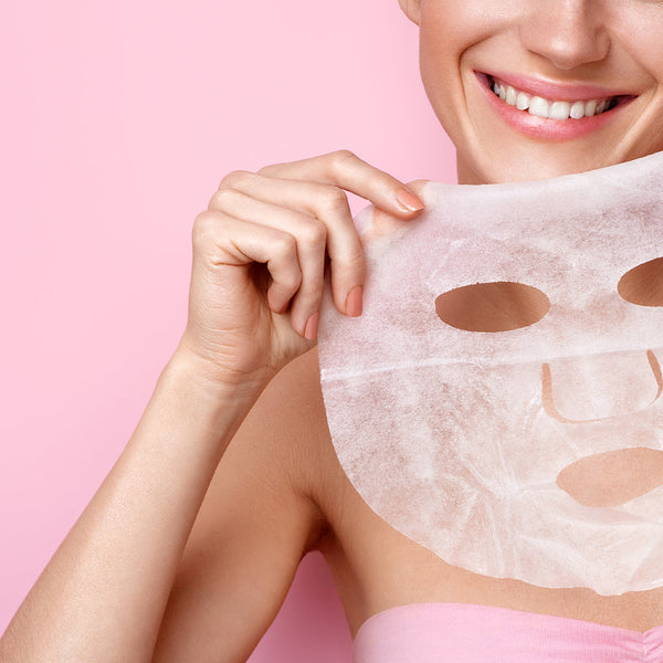 Smiling woman holding a white facial sheet mask near her face against a pink background, highlighting skincare and wellness with Vita Brightening Skin Renewal Mask | Leaders.