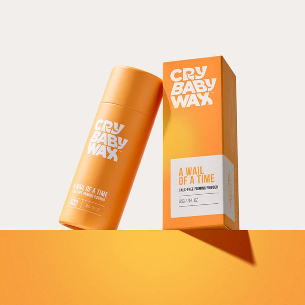 Orange cylindrical container and matching box of A Wail of a Time Priming Powder | Crybaby Wax displayed on an orange surface with light shadow, promoting skincare product.