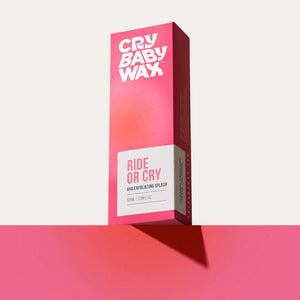 Bright pink and white packaging box of Ride or Cry AHA Exfoliating Splash | Crybaby Wax standing upright on a matching pink surface against a pale background, showcasing vibrant skincare product branding.