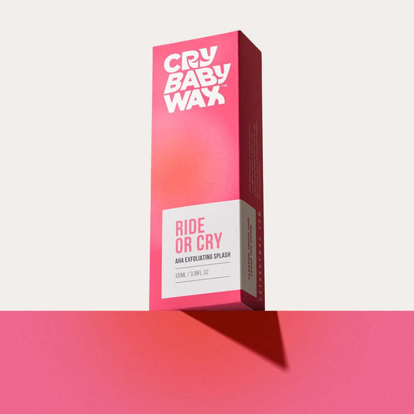 Bright pink and white packaging box of Ride or Cry AHA Exfoliating Splash | Crybaby Wax standing upright on a matching pink surface against a pale background, showcasing vibrant skincare product branding.