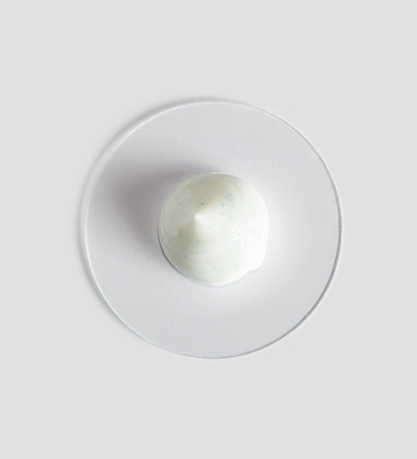 A close-up of a small amount of creamy white exfoliating scrub with fine green particles, displayed on a transparent surface, representing Essential Scrub | [ comfort zone ] skincare product.