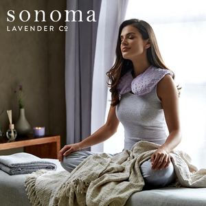 Woman wearing a textured lavender-colored Spa Mask - Lavender | Sonoma Lavender around her neck, sitting relaxed on a bed with cozy blankets in a softly lit wellness space.