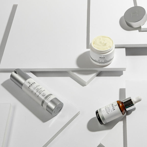 Three IMAGE Skincare products on white geometric surfaces, featuring AGELESS total anti-aging serum | IMAGE Skincare with a dropper bottle, a cream jar, and a silver pump bottle for skincare and wellness.
