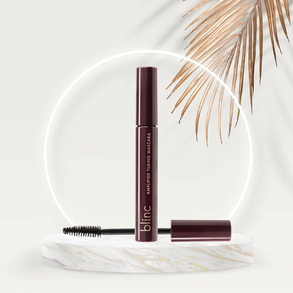 Dark burgundy tube of Amplified Tubing Mascara | Blinc with the mascara wand resting on a white marble platform, surrounded by a soft glowing ring and dried palm leaf accents.