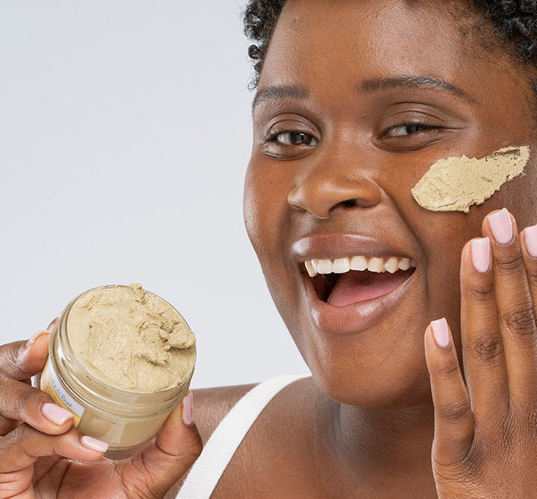 Smiling woman applying the Matcha Purity™ Calm & Clear Purification Mask | FarmHouse Fresh to her cheek, holding an open jar of creamy green skincare mask for calming and clear skin.