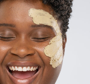 Close-up of a smiling person with eyes closed applying Matcha Purity™ Calm & Clear Purification Mask | FarmHouse Fresh on their cheek, showcasing skincare and wellness.