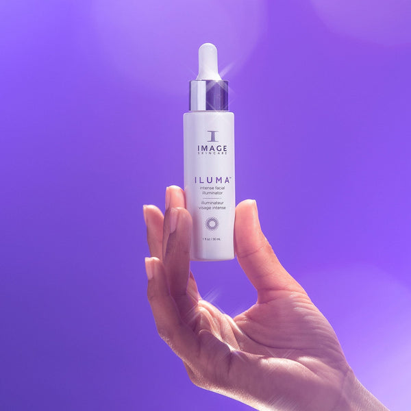 Hand holding a white dropper bottle of ILUMA intense facial illuminator | IMAGE Skincare against a purple background, showcasing skincare product for radiant skin.