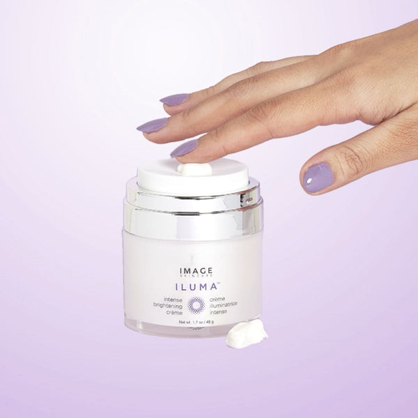 A hand with purple nail polish gently presses the pump of ILUMA intense brightening crème | IMAGE Skincare on a pastel lavender background, with a small amount of cream visible.