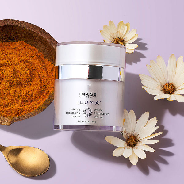 ILUMA intense brightening crème | IMAGE Skincare jar with a silver lid, placed on a lavender surface next to white flowers, a wooden bowl of orange powder, and a gold spoon.