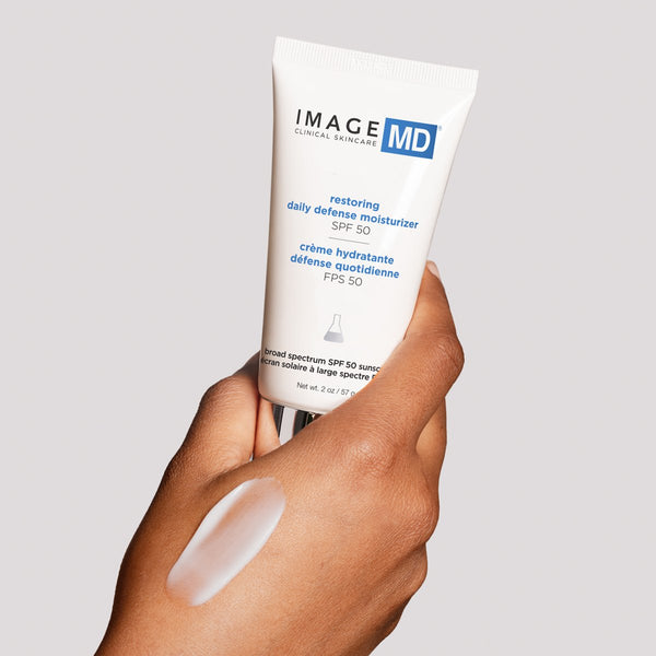 Hand holding a tube of Restoring Daily Defense Moisturizer SPF 50 | IMAGE Skincare with a sample of moisturizer applied on the back of the hand against a neutral background.