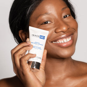 Smiling woman holding a tube of Restoring Daily Defense Moisturizer SPF 50 | IMAGE Skincare, showcasing skincare and sun protection product for healthy skin.