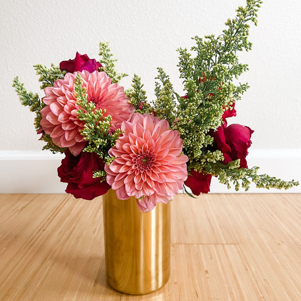 Gold Flower Vase - Live Love Spa Exclusive holding a vibrant bouquet with pink dahlias, deep red roses, and green foliage on a wooden surface against a white wall.