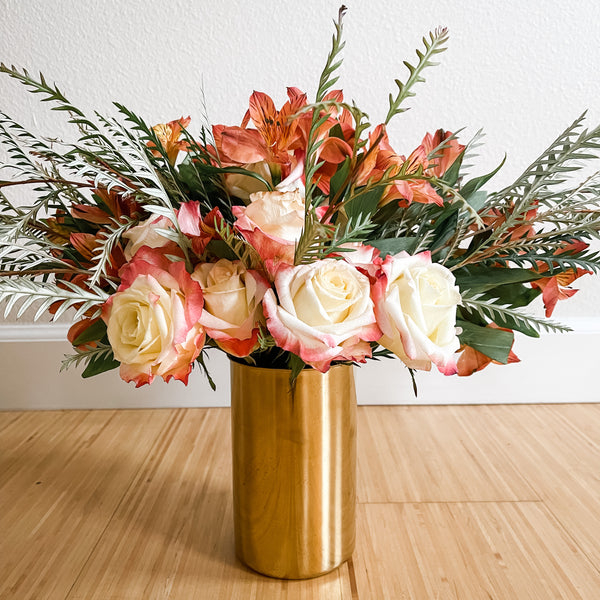 Elegant bouquet of cream and pink roses with orange flowers and greenery arranged in a sleek gold vase, Gold Flower Vase - Live Love Spa Exclusive, on a wooden floor.