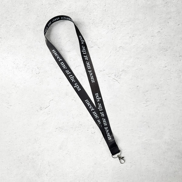 Black lanyard with white text reading "meet me at the spa" and a metal clasp, styled for skincare and wellness accessories, named Lanyard - Black Meet Me at the Spa | Lucky Owl.