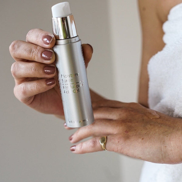 Person holding a silver bottle of Moisture Surge Hyaluronic Acid Gel | Epicuren Discovery, applying gel on the back of their hand, showcasing skincare use.