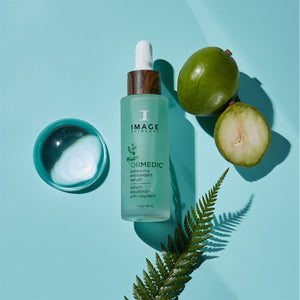 ORMEDIC balancing antioxidant serum | IMAGE Skincare bottle with wooden cap next to a small bowl of clear liquid, green fruit halves, and a fern leaf on a light blue background.
