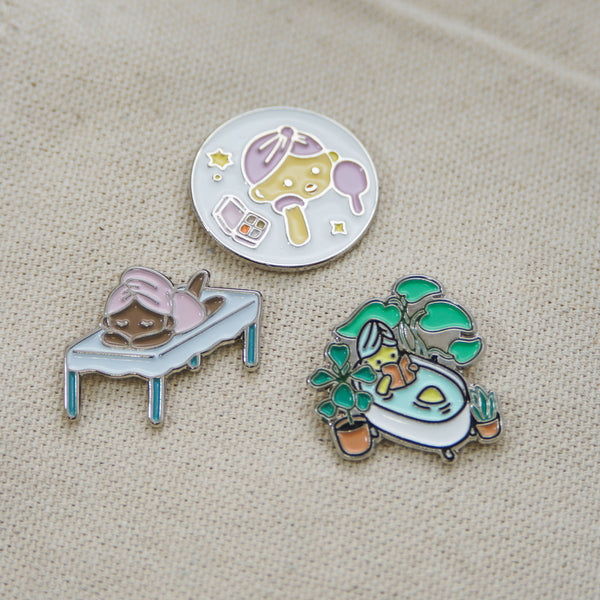 Three colorful enamel pins featuring spa and wellness themes including facial care, relaxation on a massage table, and a cozy bathtub surrounded by plants, showcasing the Fab Spa Life Enamel Pin - Limited Edition | Lucky Owl.