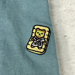Close-up of a light blue fabric patch featuring a bear in a spa robe lounging on a mat, embroidered on the Lounging Spa Bear Comfy Tee | Lucky Owl.