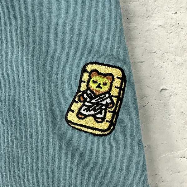Close-up of a light blue fabric patch featuring a bear in a spa robe lounging on a mat, embroidered on the Lounging Spa Bear Comfy Tee | Lucky Owl.