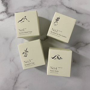 Four cream-colored candle boxes arranged on a marble surface, featuring minimalist black illustrations and text, highlighting No. 1 Sunrise Island Yoga (Bali) Candle | Lucky Owl.
