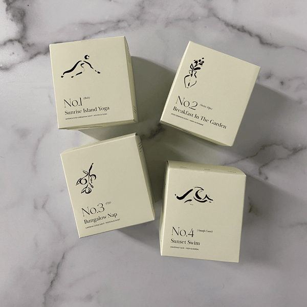Four cream-colored candle boxes arranged on a marble surface, featuring minimalist black illustrations and text, highlighting No. 1 Sunrise Island Yoga (Bali) Candle | Lucky Owl.