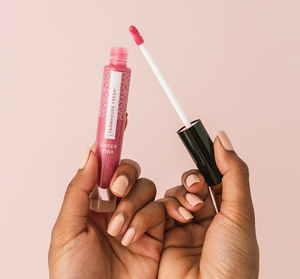 Hand holding an open tube of Vitamin Glaze® Oil Infused Lip Gloss – Sheer Pink | Farmhouse Fresh with applicator showing glossy pink lip color against a soft pink background.