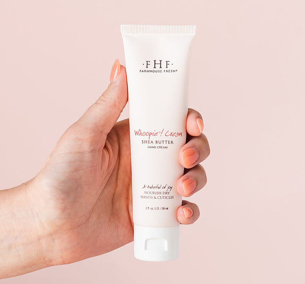 Hand holding a tube of Whoopie® Cream Shea Butter Hand Cream | Farmhouse Fresh against a soft pink background, showcasing skincare product for nourishing dry hands and cuticles.