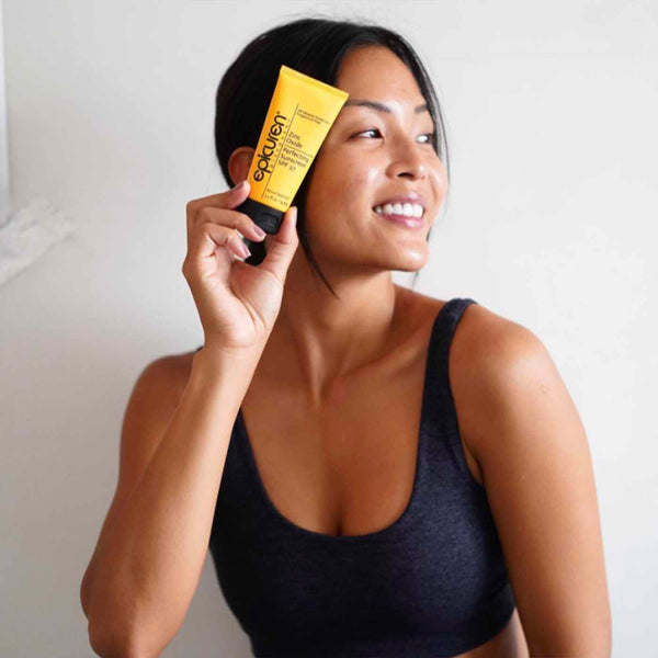 A smiling woman in a dark sports bra holds a yellow tube of Zinc Oxide Perfecting Sunscreen | Epicuren Discovery near her face, promoting skincare and sun protection.