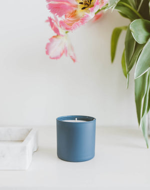 Oakmoss + Amber Dignity Series Glass Jar Soy Candle | Calyan Wax Co. - 5.3 oz sitting on a white surface next to a marble tray with pink flowers and green leaves in the background.