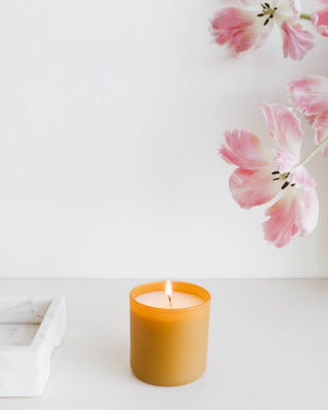Lit amber glass candle on white surface near pink tulip flowers with white wall background, featuring Vetiver + Tonka Dignity Series Glass Jar Soy Candle | Calyan Wax Co. - 5.3 oz